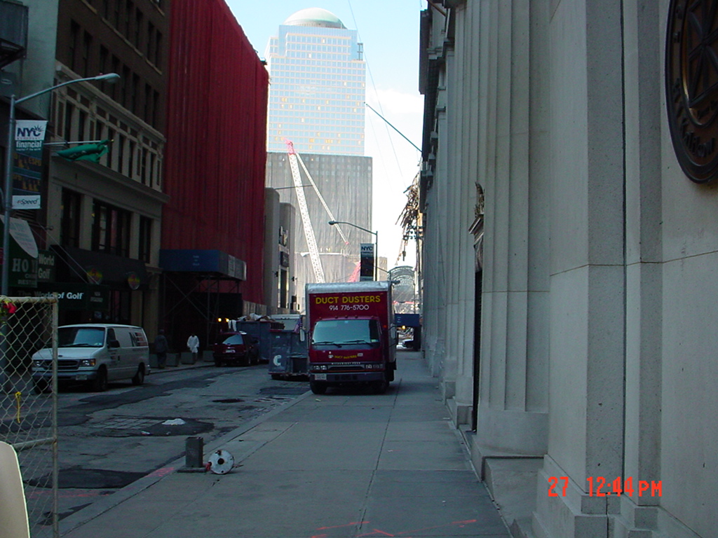 A Lower Manhattan street with construction fencing, trucks, cranes, and the World Financial Center visible beyond the cleanup area.