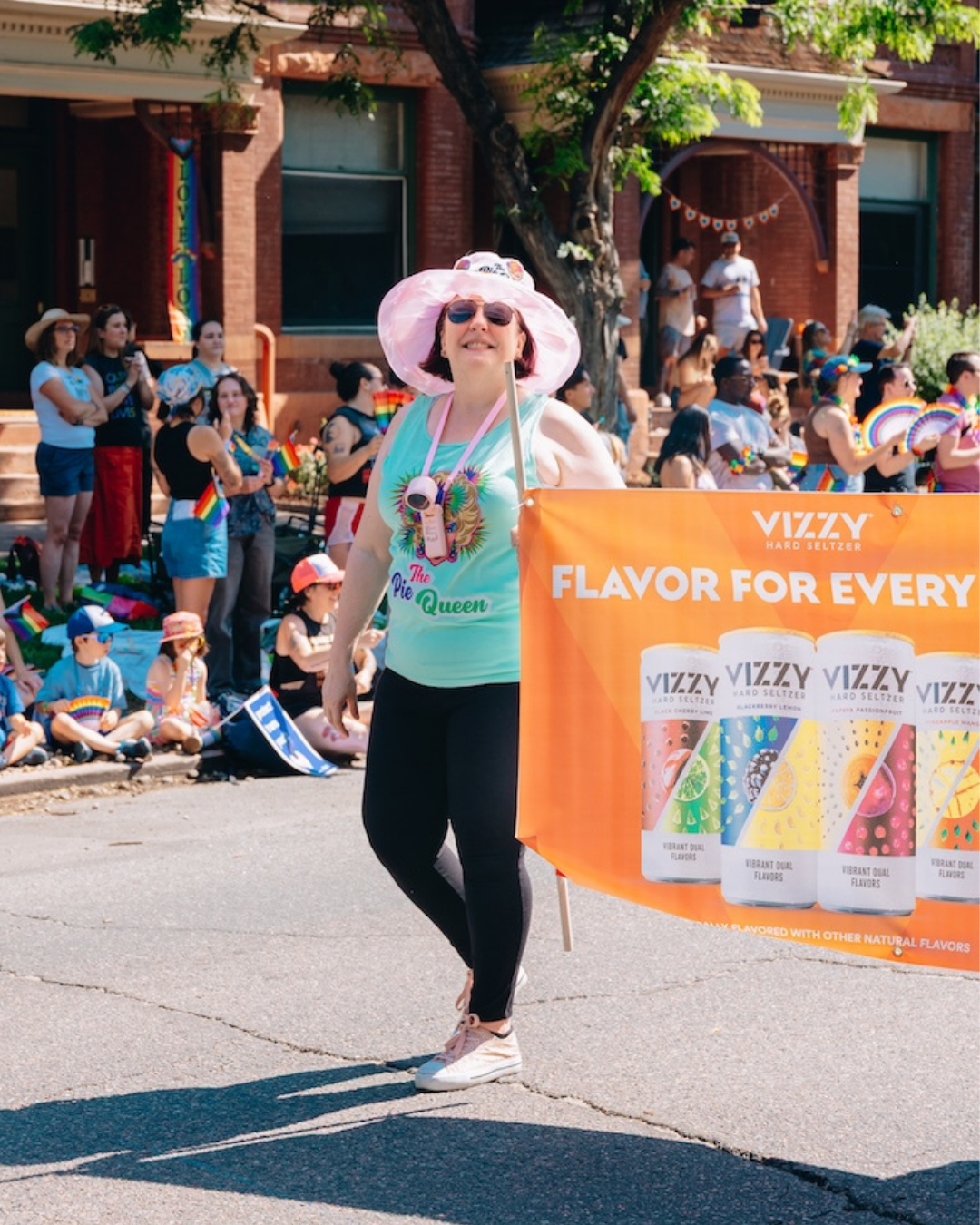 Lavinia Draper marching in the Denver Pride Parade, pink bucket hat and Pie Queen shirt, carrying a Vizzy Hard Seltzer banner.